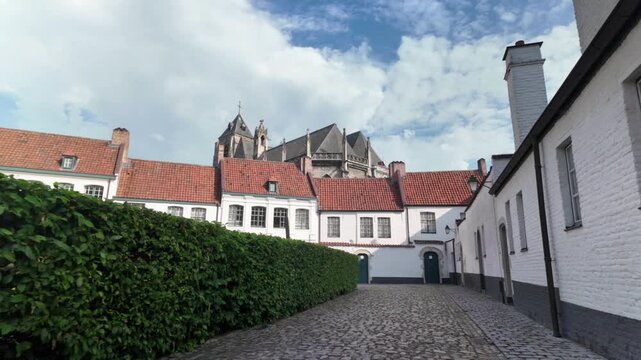 Historic UNESCO Courtrai beguinage forward walk revealing white heritage houses and the towering Gothic church in soft daylight.