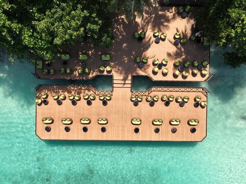 Aerial view of a wooden deck with green lounge chairs over turquoise water surrounded by lush tropical trees Maldives.