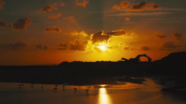 Sunset over a tranquil beach with silhouetted clouds and machinery, reflecting golden light on calm water, creating a serene coastal atmosphere at dusk