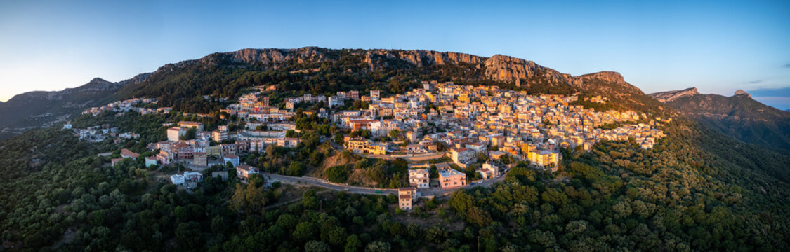 Aerial view of the panoramic historic town of Baunei perched on a rugged mountain slope with colorful houses, dense green forests, and rocky cliffs in Baunei, Sardinia, Italy.