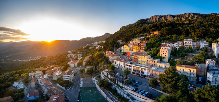 Aerial view of the hillside town of Baunei featuring colorful buildings and a football pitch during a golden sunset over the mountains in Baunei, Sardinia, Italy.