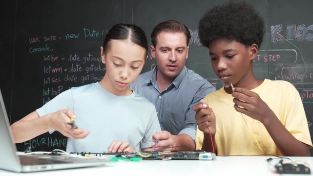Caucasian teacher talking about electric tool while student fixing robotic model on table with electronic equipment placed. Diverse academic children learning about main board structure. Edification