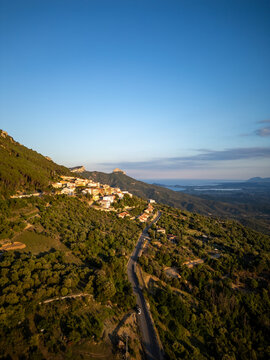 Aerial view of the mountain village of Baunei perched on a steep hillside with lush green forests, a winding road, and the Mediterranean Sea in Baunei, Sardinia, Italy.