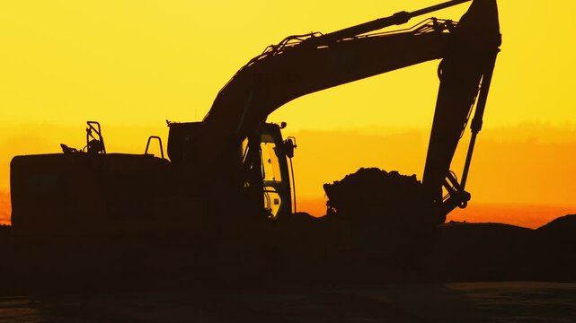 Excavator lifting and dumping soil at sunset, showcasing the continuous action of earthmoving in a construction site with a vibrant orange sky