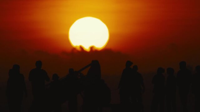 Silhouetted figures gather on a beach at sunset, enjoying the warm glow of the sun as it descends on the horizon, creating a serene atmosphere