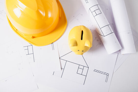 Construction plans with yellow helmet and piggy bank on table, closeup