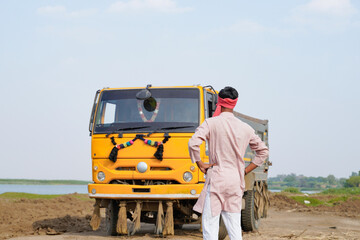 Indian truck driver standing confidently from the back beside a yellow dumper truck on a rural construction road © Niks Ads