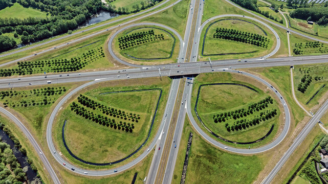 Aerial view of a cloverleaf highway interchange with symmetrical loops, rows of planted trees, and vehicles traveling along the asphalt roads in Heerenveen, Friesland, Netherlands.