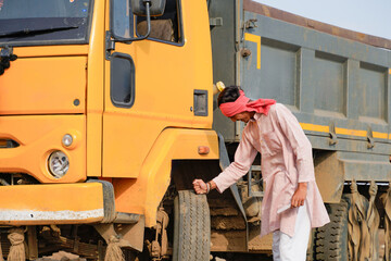 Smiling Indian truck driver in traditional attire posing confidently beside a yellow dumper truck in a rural outdoor location. © Niks Ads