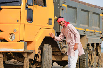 Smiling Indian truck driver in traditional attire posing confidently beside a yellow dumper truck in a rural outdoor location. © Niks Ads