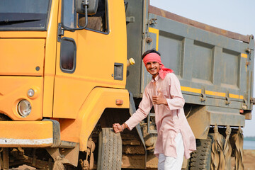 Smiling Indian truck driver in traditional attire posing confidently beside a yellow dumper truck in a rural outdoor location. © Niks Ads