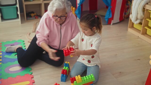 Woman hands a toy block with her hand to a girl while sitting on a foam mat in a building playroom; intergenerational bonding tenderness.