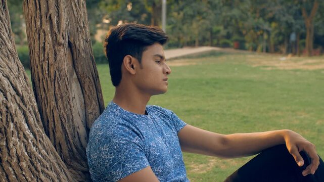 Upset young man thinking about his life while sitting near a tree trunk in the park. A concerned youngster wearing a printed T-shirt feeling lonely and sad while sitting on the garden ground - anxi...