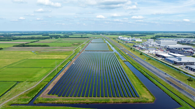 Aerial view of a large solar farm with rows of photovoltaic panels surrounded by canals and green fields near an industrial area and highway Heerenveen, Friesland, Netherlands.