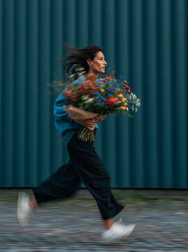 Young Woman Running Through Urban Street Carrying Colorful Wildflower Bouquet With Motion Blur Background in Blue Jacket