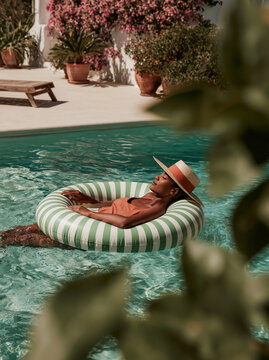 Young Woman Relaxing in Inflatable Ring Floating in Blue Swimming Pool With Straw Hat in Lush Mediterranean Summer Resort Setting