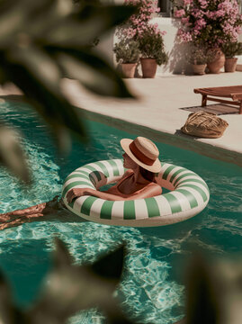 Young Woman Relaxing in Inflatable Ring Floating in Blue Swimming Pool With Straw Hat in Lush Mediterranean Summer Resort Setting