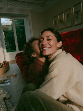 Happy Mother and Child Laughing Together on Red Couch in Cozy Sunlit Living Room With Warm Home Interior and Natural Window Light
