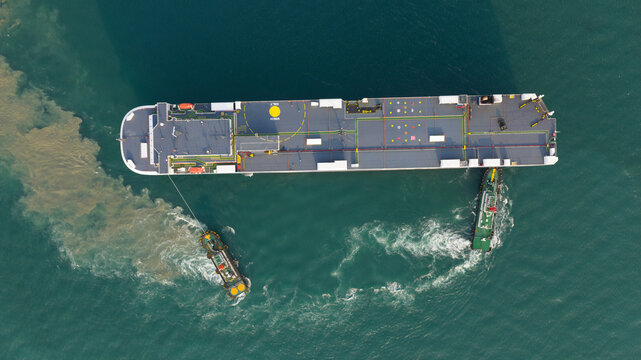 Aerial view of a large cargo ship being assisted by two tugboats in the turquoise waters of the port Laem Chabang, Chon Buri, Thailand.