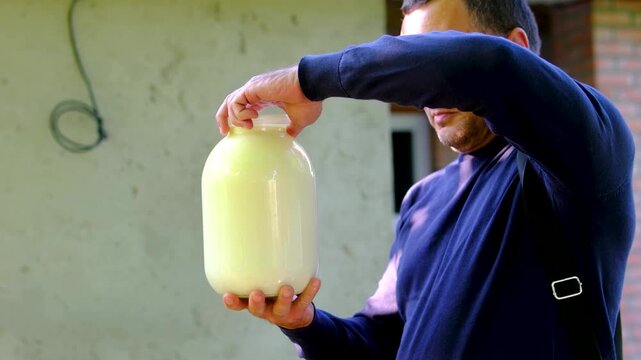 A man stands outdoors near a village house holding a large three liter jar of fresh homemade milk. Traditional countryside lifestyle, farm products and local dairy food.