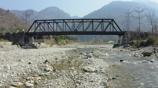 Aerial drone footage of a steel truss bridge spanning a rocky riverbed surrounded by scenic Himalayan foothills near Dehradun, Uttarakhand, India. Natural mountain landscape with flowing stream, count
