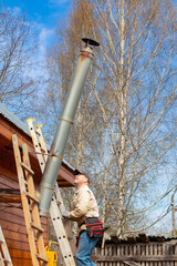 A man on a ladder inspecting a high metal chimney pipe of a wooden country house. Checking the heating system and home maintenance concept. © Chebotaeva Ekaterina