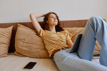 Fototapeta premium A relaxed woman lounges on a couch at home with a laptop on her lap, a smartphone nearby. A casual lifestyle moment highlighting comfort, downtime, and home work balance.