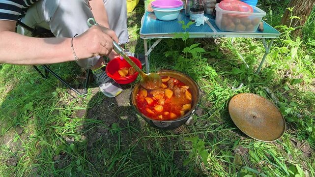 Woman scooping hot goulash soup from cauldron into bowl during outdoor picnic