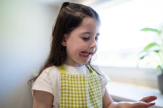 Child in apron tasting sweet dough from finger indoors