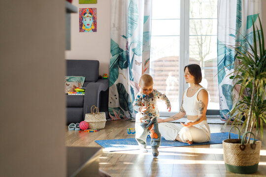 Woman meditating on yoga mat in living room while baby boy walks