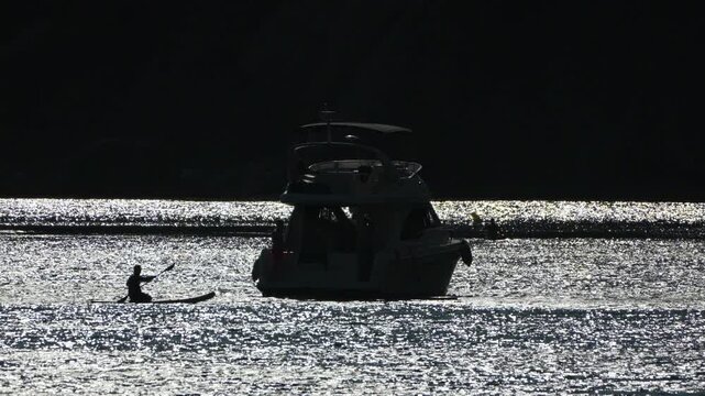 Paddleboard yacht silhouette person on a standup board glides past a luxury boat on shimmering water during a tranquil evening