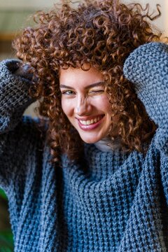 Smiling woman winking while holding curly hair in knit sweater indoors