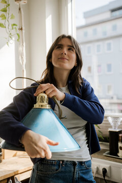 Young woman holding a new lamp by the window while planning decoration