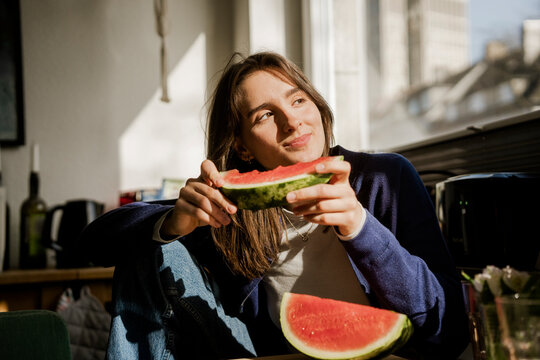 Smiling woman eating fresh watermelon by the window at home