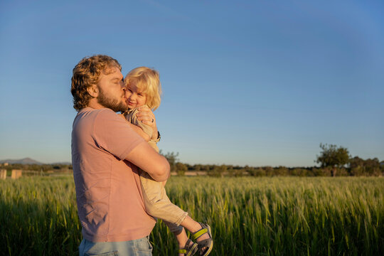 Happy father holding and kissing kid in summer field at sunset