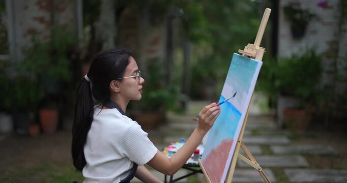 asian female artist painting horizon line on canvas sitting in garden holding brush with precision creating soft gradient between sky and sea during quiet creative outdoor moment