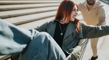 Obraz premium A young red-haired woman laughs while sitting in a shopping cart being pushed by a male friend. They enjoy a playful outdoor moment on steps as she stretches her arms and smiles