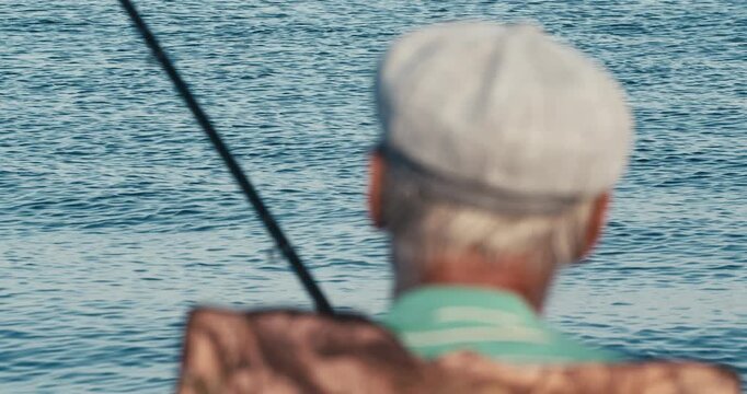 An elderly man in a flat cap fishes by the calm sea, seen from behind with his rod in hand.