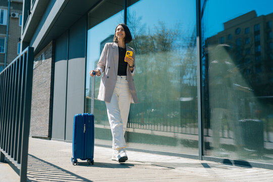 Smiling businesswoman walking outdoors with suitcase and smartphone