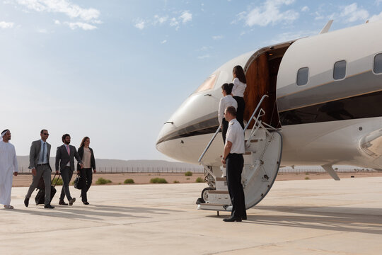 Smartly dressed business travelers with traditional Gulf Arab partner approach a luxury private jet as elegant flight crew welcome them aboard on a sunny desert airport tarmac ideal for premium