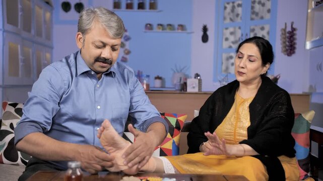Caring Indian husband putting a cloth / crepe bandage on his injured wife's feet. Medium shot of an attractive housewife with a wounded leg on a table giving painful expression - health and medical
