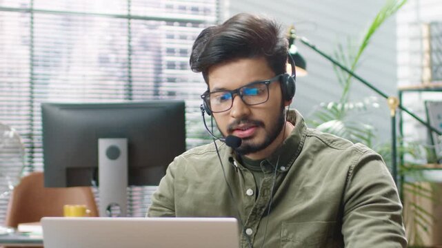 Man wearing headset and speaking during video call. Sitting by laptop in bright office. Male customer support agent answering online query, engaged in remote conversation in modern workspace.