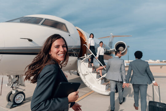 Professional young businesswoman in navy blazer holding digital tablet greets arriving business executives with warm smile at white private corporate jet aircraft on sunny coastal airport tarmac with
