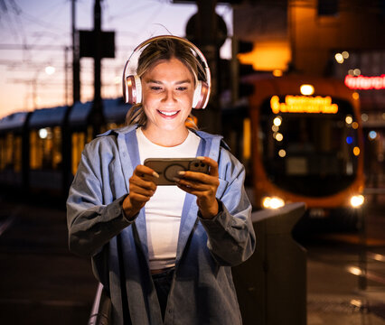 Woman smiling at smartphone with headphone in city at night