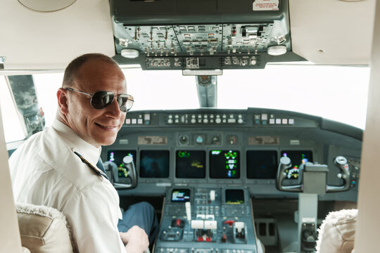 Confident professional airline pilot wearing aviator sunglasses and white shirt smiling over his shoulder inside a modern aircraft cockpit with illuminated avionics displays ideal for aviation and