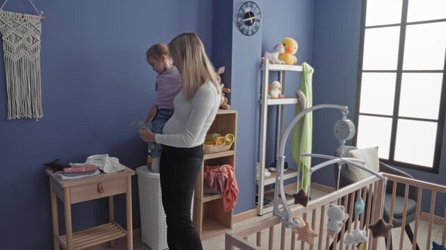 Woman holding toddler at wooden crib, hands supporting child while handing toy near a large window in a nursery building; tenderness bonding.