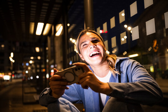 Smiling commuter playing mobile game at city bus stop at night