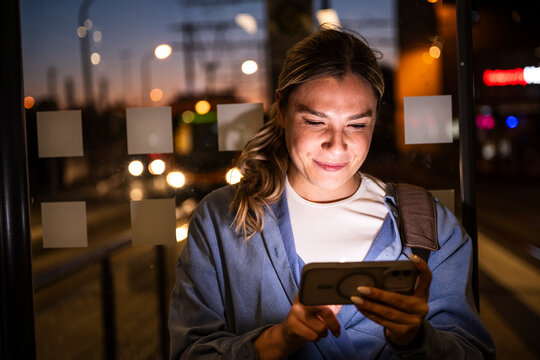 Female commuter smiling while using mobile phone at bus stop at night