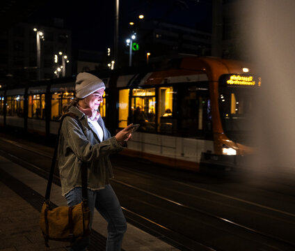 Smiling woman using smartphone at city tram station at night