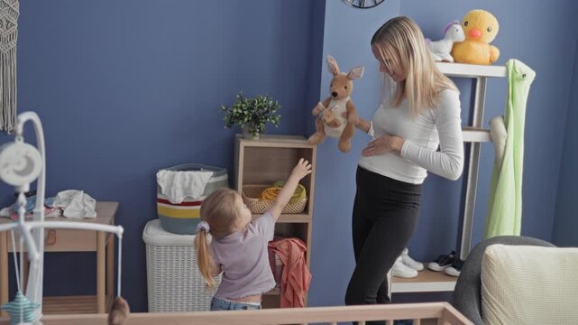 Woman handing stuffed kangaroo to little daughter, child reaching with small hand while mother touches belly beside crib and mobile in nursery building; tenderness bonding.